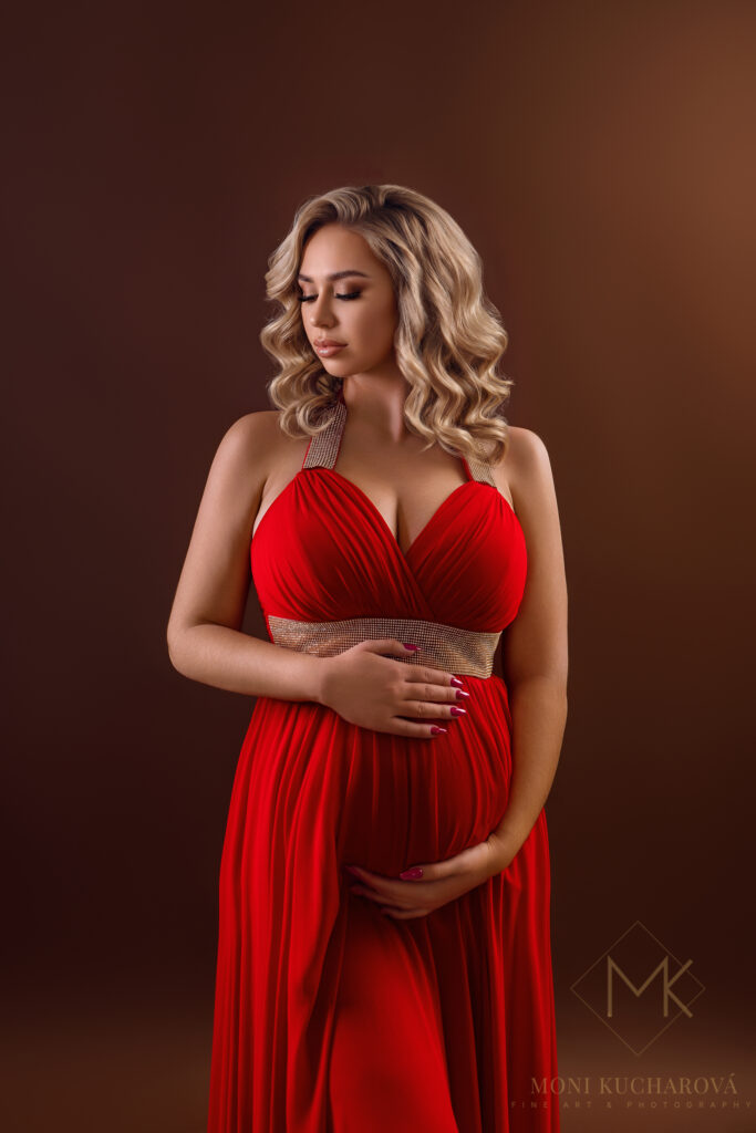 Ukážka luxusného tehotenského fotenia v Bratislave. Krásna tehotná žena v elegantných červených šatách na tmavom pozadí. Profesionálny fine art portrét vytvorený fotografkou Moni Kucharovou.
